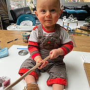 Gabriel participe au concours pour gagner de l'argent avec cette photo : toddler, child, baby, indoor, overalls, striped_clothing, wooden_stick, table, shoes, face, blue_eyes, living_room, furniture, cup, blanket, high_chair, plant, toy, window, floor