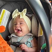 Aaliyah is registered to the contest to win money with this photo: baby, car_seat, headband, bow, toy, orange, smiling, happy, infant, child, safety_strap, clothing, seatbelt, cute, indoors, hand, face, person, car_interior, playful