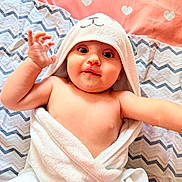 Luis is registered to the contest to win money with this photo: baby, infant, child, portrait, smiling, waving, hooded_towel, bathrobe, blanket, bedding, heart_pattern, bed, lying_down, indoor, close_up, face, eyes, hands, cozy, photography
