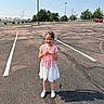child, girl, smiling, outdoor, parking_lot, parking_spaces, dress, pink_clothing, floral_dress, hair_bows, sandals, socks, standing, portrait, sunlight, shadow, asphalt, trees, cars, sky