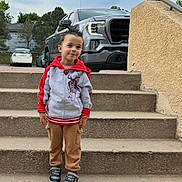 Juan is registered to the contest to win money with this photo: child, young_boy, smiling, outdoor, portrait, standing, stairs, driveway, pickup_truck, vehicles, suburban, house, trees, sky, jacket, hoodie, pants, sneakers, concrete, daylight