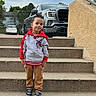 child, young_boy, smiling, outdoor, portrait, standing, stairs, driveway, pickup_truck, vehicles, suburban, house, trees, sky, jacket, hoodie, pants, sneakers, concrete, daylight