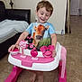 child, baby_walker, toy, tshirt, cow_print, floor, tile, couch, lamp, indoor, happy, smile, person, feet, furniture, play, young_child, home, walking_aid, pink