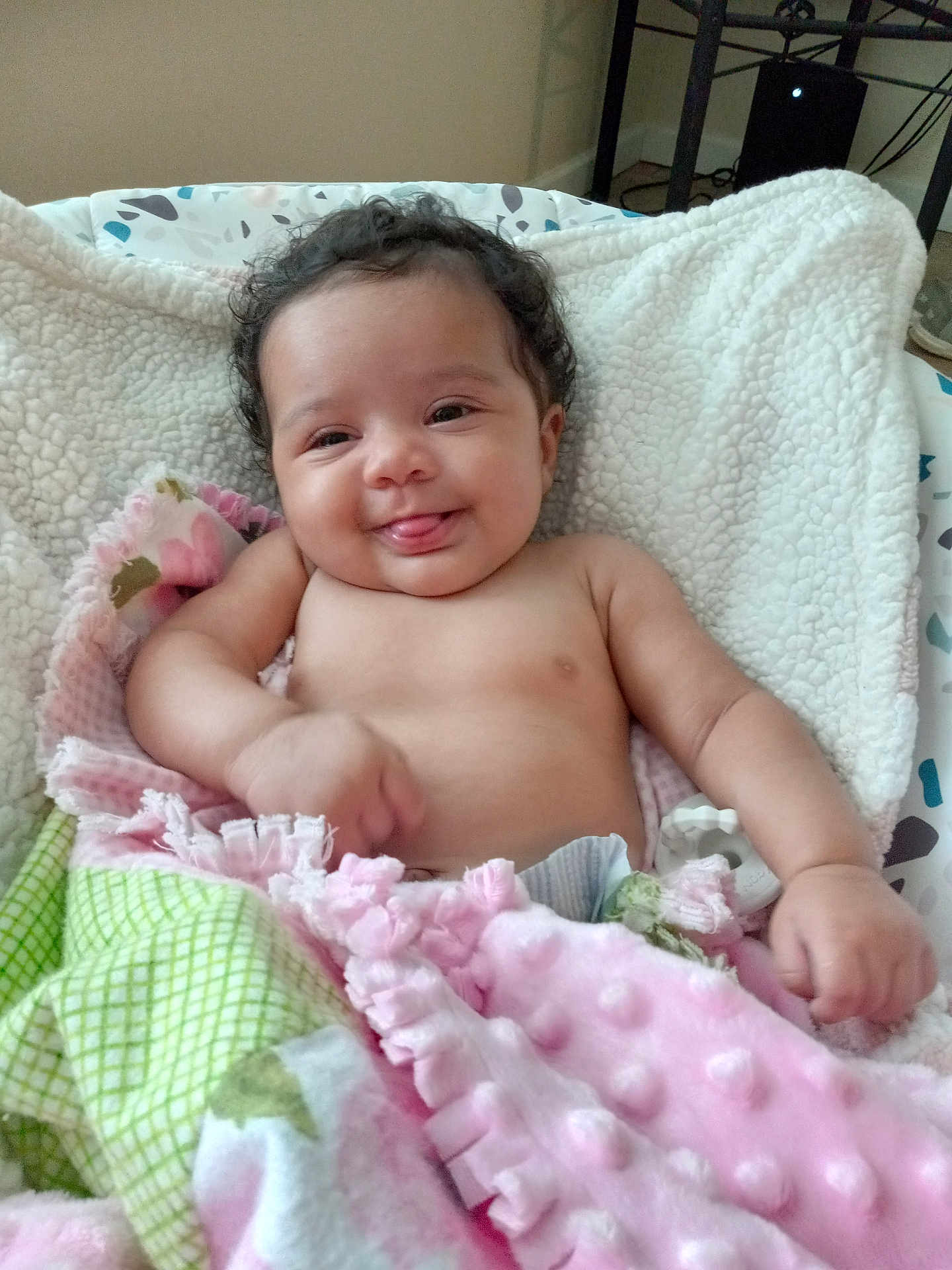 Beretta is registered to the contest to win money with this photo: baby, smiling, tongue_out, blanket, curly_hair, indoor, infant, cute, child, cozy, soft_texture, pink, green, white, happy, person, lying_down, closeup, warm, comfort
