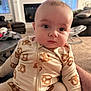 baby, infant, onesie, pretzel_pattern, indoor, living_room, carpet, sofa, table, face, person, child, cute, seated, clothing, home, expression, curious, soft_light, portrait