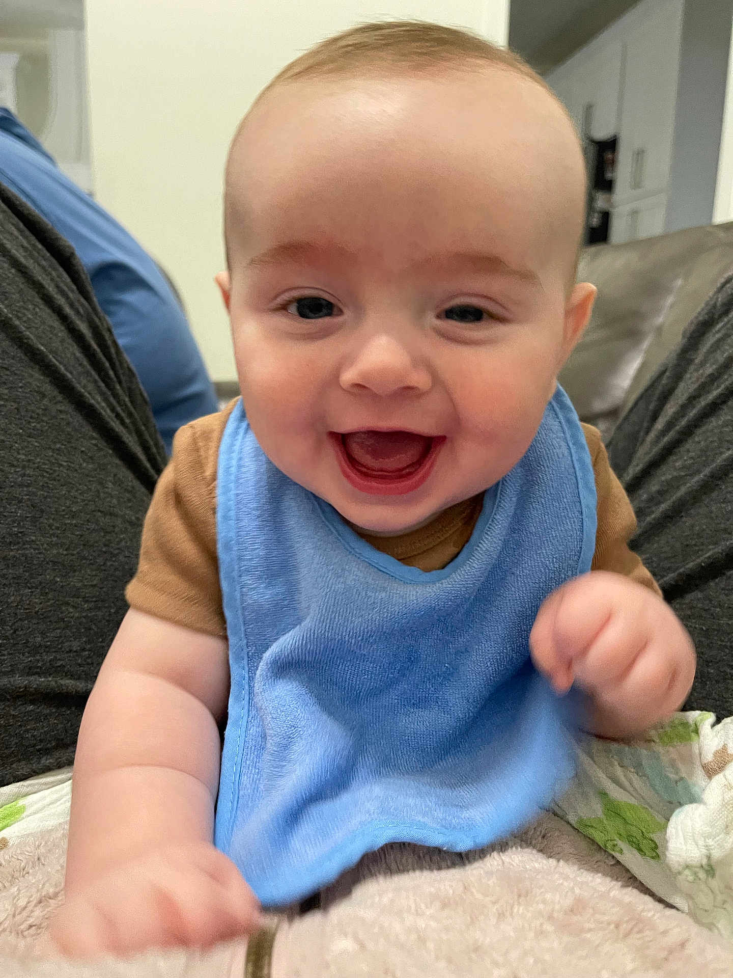 Ash joined the competition — help win amazing prizes! baby, smiling, blue_bib, brown_shirt, happy, infant, face, person, hand, home_interior, couch, blanket, seated, cute, child, young, skin, expression, portrait, indoor