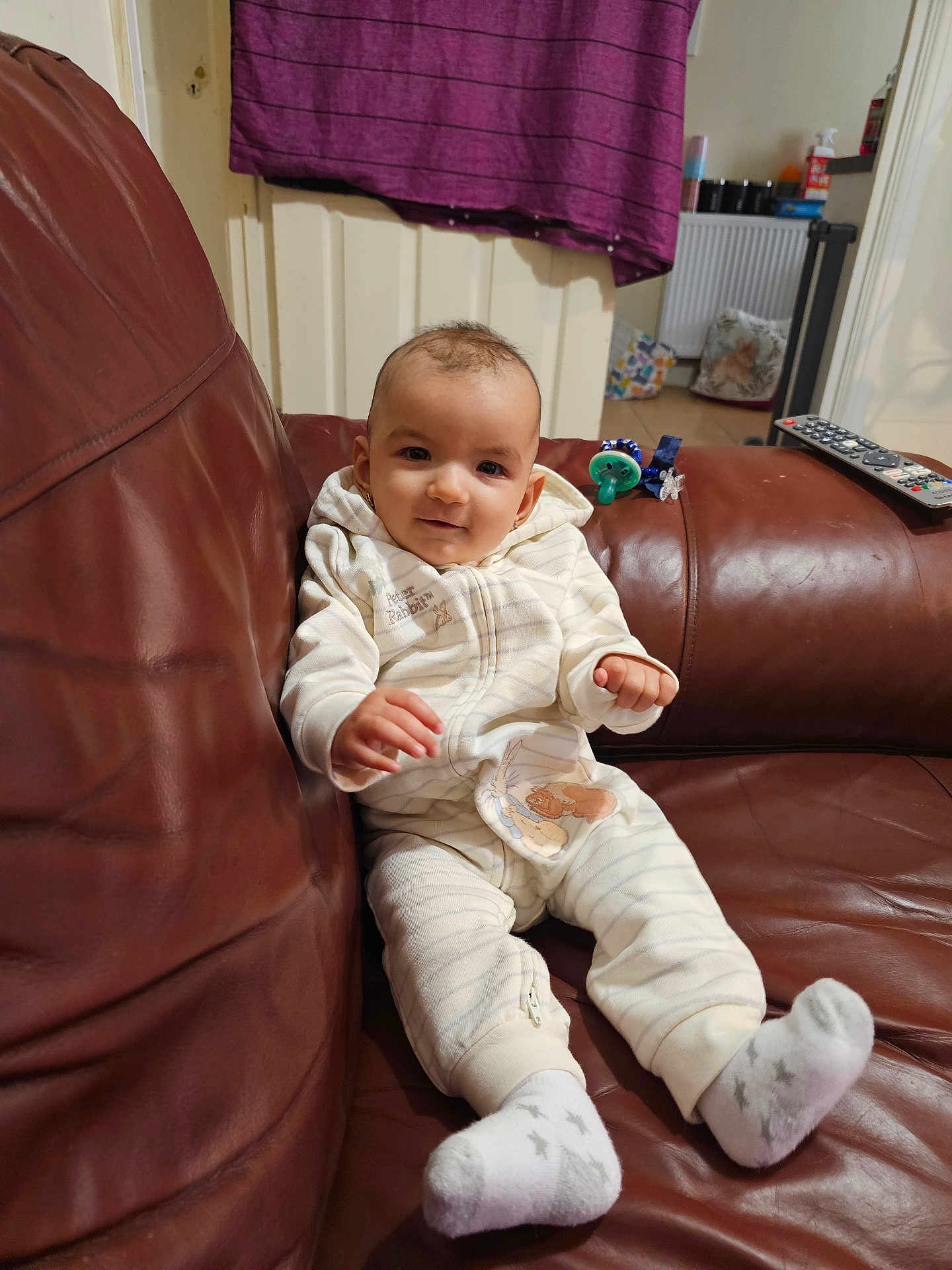 Annika is registered to the contest to win money with this photo: baby, child, couch, leather, onesie, socks, remote_control, door, towel, indoor, furniture, person, face, expression, cozy, casual, relaxed, home, infant, curious