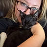 person, cat, glasses, black_cat, white_paws, cuddle, sleeping_cat, indoor, close_up, arm, face, relaxation, comfort, leather_chair, blonde_hair, casual_clothing, home, pet, affection, portrait