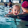 baby, child, swimming_pool, water, adult, swim_cap, blue_eyes, splashing, happy, smiling, person, swimming, lesson, indoor, recreation, fun, support, blurred_background, face, head