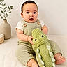 baby, child, plush_toy, crocodile, green_overalls, white_shirt, sitting, indoors, vase, plant, decor, neutral_background, barefoot, cute, infant, portrait, soft_toy, smiling, texture, casual_clothing
