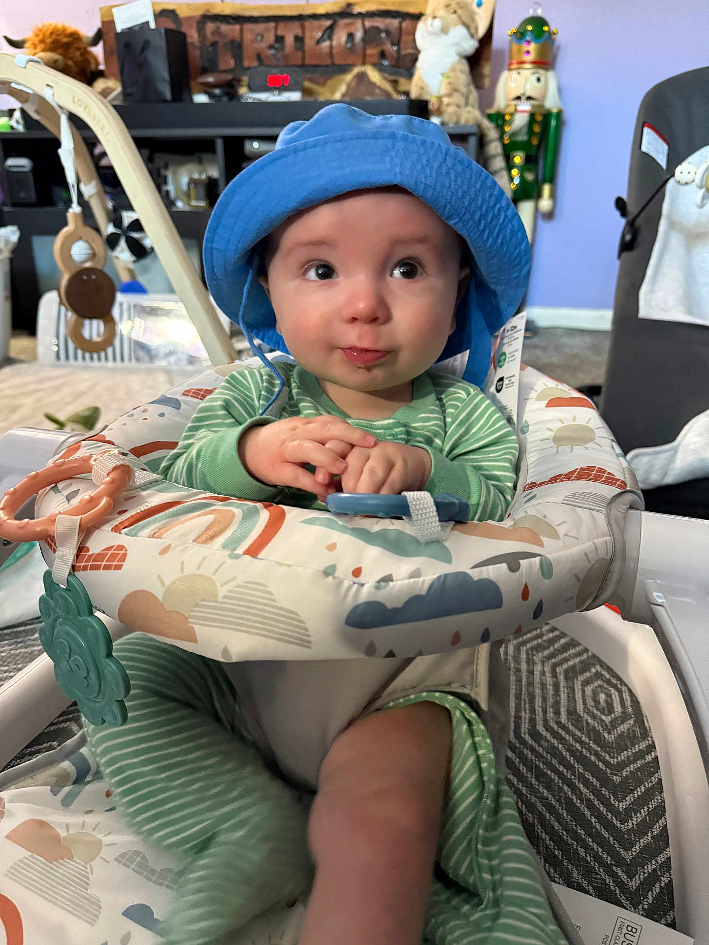 Silas is registered to the contest to win money with this photo: baby, blue_hat, green_pajamas, baby_seat, toy, indoor, child, cute, infant, playtime, smiling, face, hands, carpet, decor, nutcracker, plush_toy, furniture, expression, person