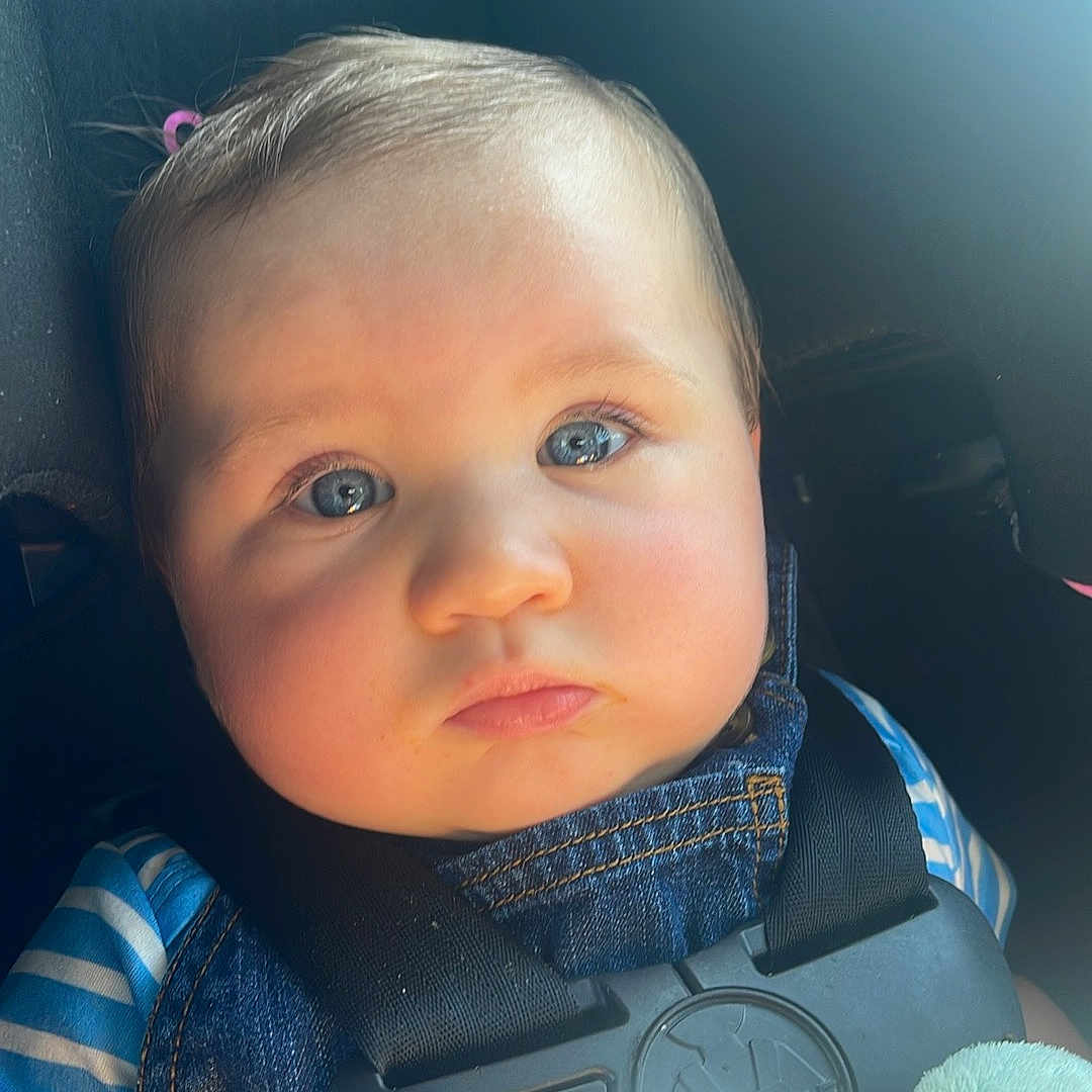 Ashton is registered to the contest to win money with this photo: baby, child, car_seat, blue_eyes, denim_overalls, striped_shirt, seatbelt, plush_toy, infant, closeup, face, portrait, safety, child_safety, cute, indoors, natural_light, expressionless, young_child, car_interior