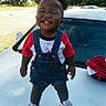 baby, car, car_hood, child, denim, facial_expression, fashion, outdoor, overalls, parked_cars, playful, portrait, smiling, sneakers, squinting, standing, sunlight, teeth, toddler, trees