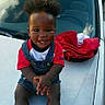 automobile, boy, car, child, curly_hair, daylight, denim_overalls, happy, hood, jacket, outdoor, photography, portrait, red_shirt, sitting, smiling, sneakers, toddler, vehicle, windshield