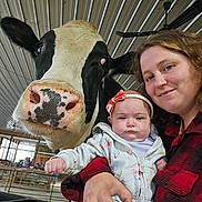 Grace is registered to the contest to win money with this photo: baby, infant, woman, cow, farm, barn, selfie, portrait, smiling, outdoor, animal, livestock, black_and_white_cow, headband, baby_clothing, holding_baby, family, barn_interior, cow_face, closeup