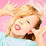 blonde_hair, blue_shirt, casual_clothing, child, close_up, cute, earrings, expression, fun, hands_up, happy, joyful, person, pink_background, playful, portrait, silly_face, studio_lighting, tongue_out, young