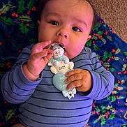 Gabriel is registered to the contest to win money with this photo: baby, infant, onesie, blue_clothing, striped_clothing, face, hand, christmas_ornament, holiday, blanket, holly_pattern, carpet, indoor, cute, smiling, person, closeup, cheeks, lying_down, festive