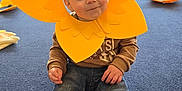Kayden is registered to the contest to win money with this photo: toddler, child, yellow_flower_headpiece, costume, indoor, blue_carpet, craft_activity, group, people, sitting, casual_clothing, jeans, brown_sweater, playful, smiling, faces, women, baby, event, fun