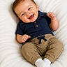 baby, child, closeup, clothing, cute, face, feet, hands, happy, infant, joy, khaki_pants, navy_blue_shirt, person, playful, portrait, sitting, smile, sock, white_blanket
