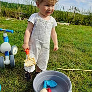 Côme a rejoint le concours — aidez-le/la à gagner de superbes lots ! bucket, child, clothing, curly_hair, daylight, footwear, garden, grass, greenery, nature, outdoor, play, rake, sand_toys, short_sleeves, smile, summer, toddler, toy, tricycle