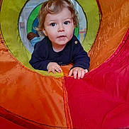 Côme participe au concours pour gagner de l'argent avec cette photo : toddler, child, play_tunnel, colorful, curious, indoor, hands, face, clothing, blue_sweater, window, background, fabric, orange, red, yellow, green, blue, person, playing