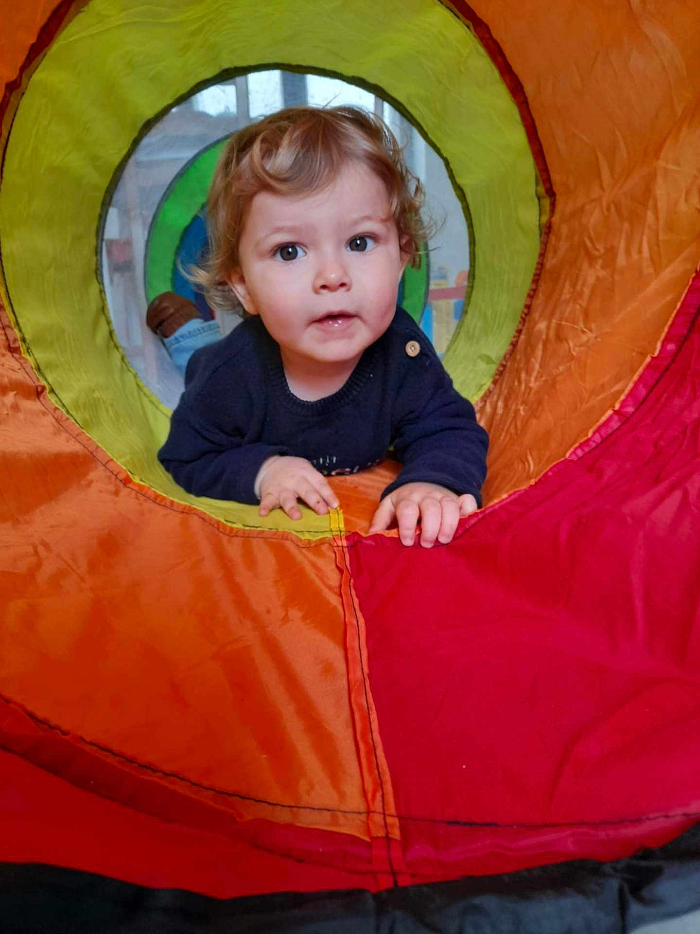 Côme participe au concours pour gagner de l'argent avec cette photo : toddler, child, play_tunnel, colorful, curious, indoor, hands, face, clothing, blue_sweater, window, background, fabric, orange, red, yellow, green, blue, person, playing