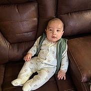 Jonathan is registered to the contest to win money with this photo: baby, infant, child, onesie, vest, brown_leather_couch, seated, indoor, cute, portrait, comfortable, quilted_fabric, patterned_clothing, small_hands, face, looking_at_camera, home_interior, soft_lighting, relaxed, young