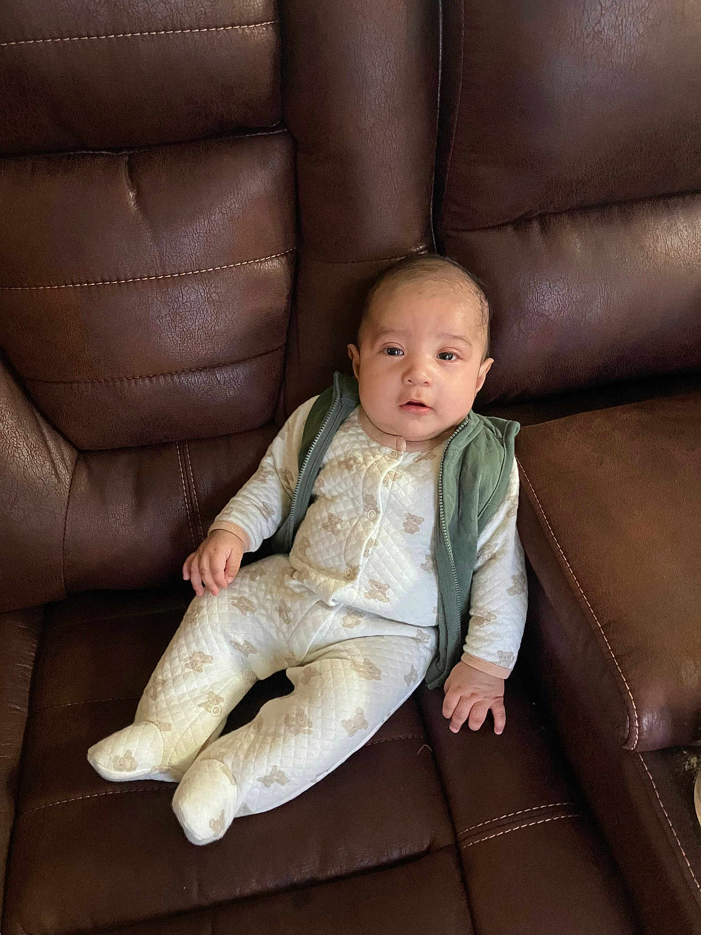 Jonathan is registered to the contest to win money with this photo: baby, infant, child, onesie, vest, brown_leather_couch, seated, indoor, cute, portrait, comfortable, quilted_fabric, patterned_clothing, small_hands, face, looking_at_camera, home_interior, soft_lighting, relaxed, young