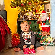 Mylan a rejoint le concours — aidez-le/la à gagner de superbes lots ! toddler, child, christmas_tree, christmas_lights, gifts, presents, holiday_decorations, santa_figurine, floor, indoor, festive, curious, sitting, red_baubles, tinsel, holiday, cozy, decor, toy, winter