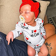Emersyn is registered to the contest to win money with this photo: baby, sleeping, pacifier, red_headband, onesie, i_love_mom, tattoo, lap, person, indoor, cushion, clothing, socks, cute, child, infant, resting, peaceful, headwear, human
