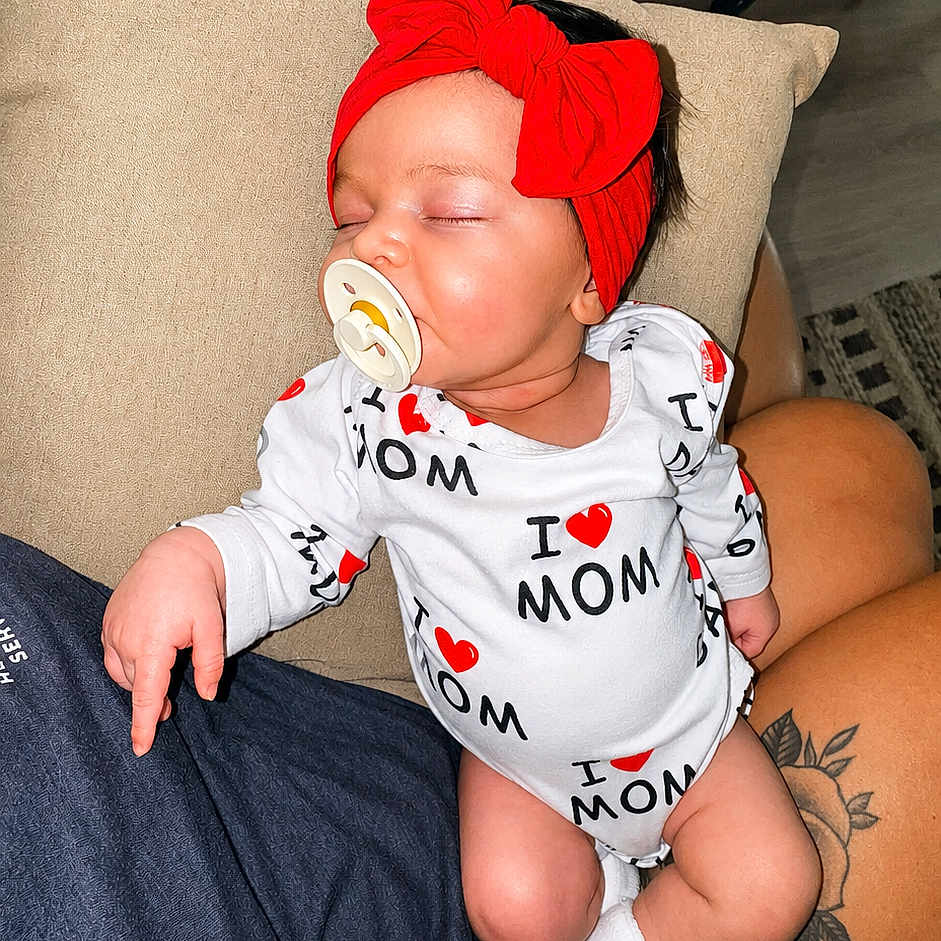 Emersyn is registered to the contest to win money with this photo: baby, sleeping, pacifier, red_headband, onesie, i_love_mom, tattoo, lap, person, indoor, cushion, clothing, socks, cute, child, infant, resting, peaceful, headwear, human