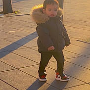 Loai participe au concours pour gagner de l'argent avec cette photo : toddler, child, puffy_jacket, hood, sneakers, shadow, sunlight, urban, pavement, outdoor, street, curious, standing, winter_clothing, person, daylight, casual, young_child, cozy, golden_hour