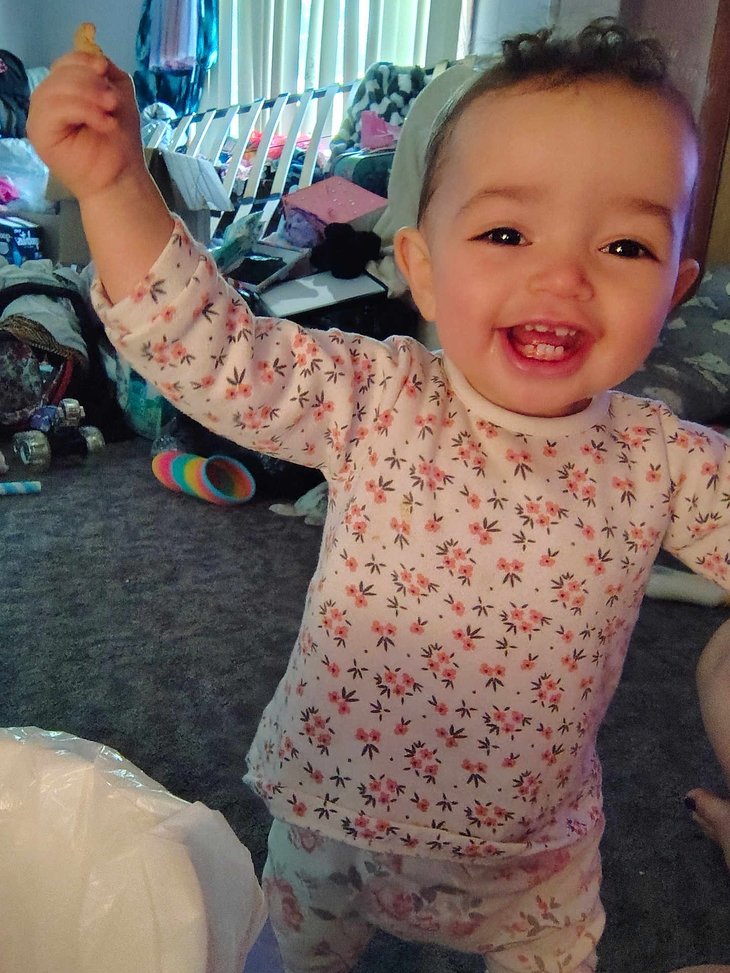Tehleyiah is registered to the contest to win money with this photo: toddler, child, smiling, happy, floral_clothing, indoors, carpet, toy, raised_arm, person, face, baby, head, living_room, window_blinds, furniture, clutter, pants, socks, curtains