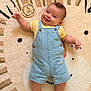 Liam participe au concours pour gagner de l'argent avec cette photo : baby, smiling, tongue_out, denim_overalls, yellow_shirt, milestone, plaque, lying_down, rug, lion_pattern, cute, infant, child, happy, chubby_cheeks, indoor, portrait, clothing, face, person