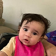 Annaliese is registered to the contest to win money with this photo: baby, casual, child, closeup, clothing, couch, curly_hair, cute, expression, face, home, indoors, infant, looking, person, pink_bib, portrait, relaxed, seated, young