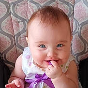 Zara joined the competition — help win amazing prizes! baby, child, face, smile, purple_ribbon, dress, patterned_cushion, highchair, happy, blue_eyes, chewing, hand, infant, cute, portrait, indoor, person, young_child, clothing, sitting