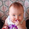 baby, child, face, smile, purple_ribbon, dress, patterned_cushion, highchair, happy, blue_eyes, chewing, hand, infant, cute, portrait, indoor, person, young_child, clothing, sitting