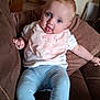 baby, child, tongue_out, blue_eyes, pink_bib, white_shirt, blue_leggings, couch, brown_furniture, home_interior, door, wooden_floor, person, infant, cute, smiling, relaxed, indoors, cozy, playful