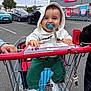 baby, child, shopping_cart, pacifier, hoodie, green_pants, white_shoes, parking_lot, supermarket, cars, outdoor, smiling, person, clothing, retail, toddler, happy, urban, daytime, cute