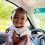 Sky is registered to the contest to win money with this photo: baby, child, smiling, hand, car, steering_wheel, interior, seat, window, daylight, person, face, hair_tie, arm, clothing, skin, automobile, joy, happy, reach