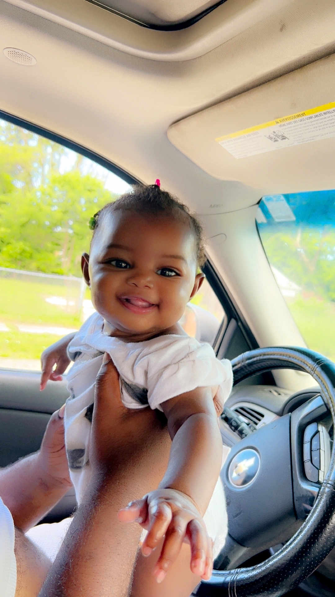 Sky is registered to the contest to win money with this photo: baby, child, smiling, hand, car, steering_wheel, interior, seat, window, daylight, person, face, hair_tie, arm, clothing, skin, automobile, joy, happy, reach