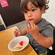 Gael is registered to the contest to win money with this photo: toddler, child, indoor, table, snack, food, disposable_bowl, watermelon, cracker, brown_hair, gray_shirt, seat, contemplative, fingers, young_child, curly_hair, face, person, eating, highchair