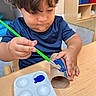 Gael is registered to the contest to win money with this photo: child, toddler, painting, paintbrush, blue_paint, palette, cardboard_tube, table, wooden_table, classroom, art_activity, short_hair, blue_shirt, indoor, focused, hand, seat, furniture, young_child, creative