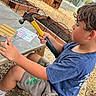 Carlos joined the competition — help win amazing prizes! child, boy, hammer, wood, bench, outdoor, garden, fence, play, tool, shorts, tshirt, person, sideview, daylight, activity, concrete, tree, woodchips, greenery