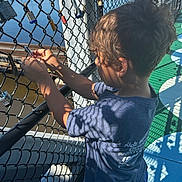 Carlos is registered to the contest to win money with this photo: child, boy, chain_link_fence, lock, sunlight, shadow, blue_shirt, outdoor, daytime, waterfront, hair, hand, fence, activity, playground, summer, casual_clothing, person, side_view, young
