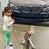Miley is registered to the contest to win money with this photo: child, cat, leash, sidewalk, wet_surface, car, damaged_car, black_car, calico_cat, girl, curly_hair, boots, green_pants, white_shirt, urban, outdoor, daytime, reflection, concrete, raindrops