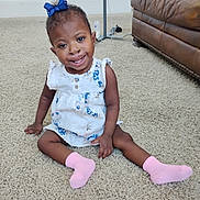 Summer is registered to the contest to win money with this photo: toddler, child, smiling, sitting, carpet, living_room, dress, pink_socks, blue_bow, happy, indoor, furniture, leather_couch, person, cute, young_child, portrait, floor, home, casual