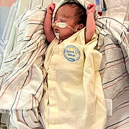 Maximillion is registered to the contest to win money with this photo: newborn, baby, infant, sleeping, blanket, hospital, crib, medical_tubes, yellow_onesie, striped_blanket, peaceful, wrapped, arms_up, soft_fabric, cute, healthcare, portrait, indoors, person, child