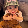 baby, child, smiling, camo_hat, overalls, pink_shirt, couch, happy, indoors, cute, face, hands, sitting, infant, clothing, person, portrait, expression, cozy, casual