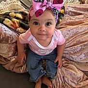 Penny is registered to the contest to win money with this photo: baby, child, pink_bow, headwear, pink_shirt, denim_pants, barefoot, bed, blanket, soft_texture, curious_expression, indoor, person, infant, cute, portrait, clothing, sitting, looking_up, face